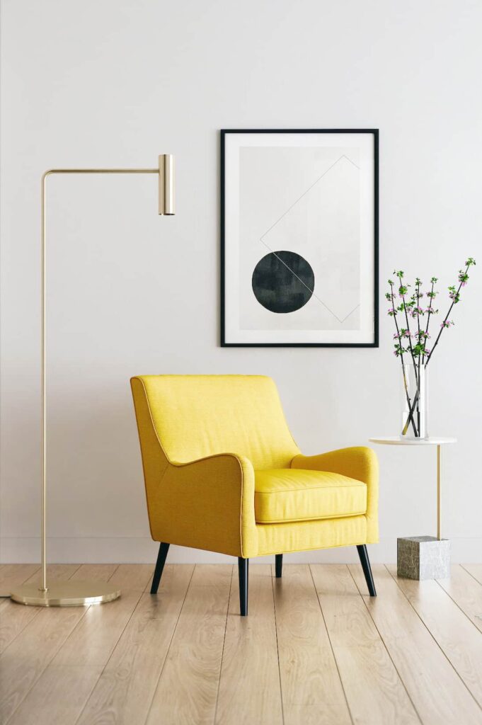 Yksitysikohta huoneesta, jossa valkoinen seinä, puulattia, keltainen matalaselkäinen nojatuoli. Nojatuolin vieressä metallinen minimalistinen jalkalamppu, toisella puolella moderni pieni pöytä, jolla oksia maljakossa. Seinällä abstrakti taulu.